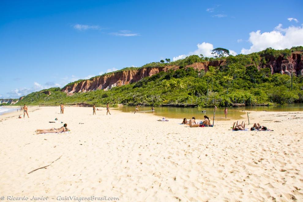 Imagem de pessoas tomando sol nas areias claras e ao fundo uma linda vegetação.